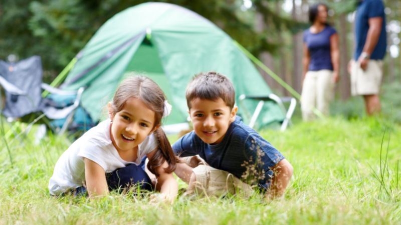 Two little kids sitting on the grass with their parents standing in the background next to a tent.