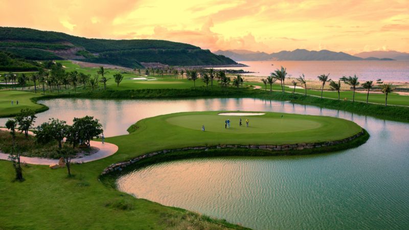 A tranquil golf course at dusk with a group of golfers standing on a green that is surrounded by water.