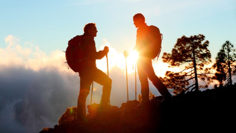 Two hikers stand on a mountain trail at sunset. They are silhouetted against the bright sun, which peeks through the trees and casts a warm glow around their figures. Both are equipped with backpacks and trekking poles, and appear to be engaged in conversation.