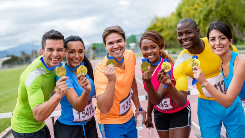 A group of six exuberant athletes displaying their medals and smiling broadly at the camera after a race, dressed in colorful running attire.