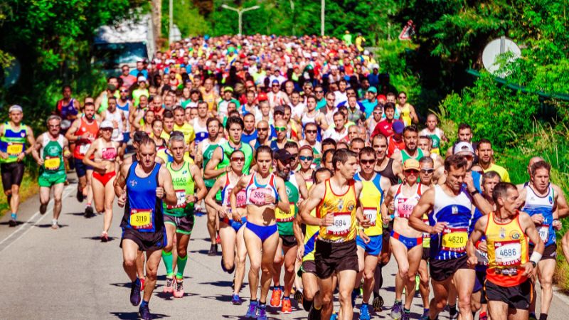 A vibrant and crowded marathon race with runners in colorful athletic gear, racing down a sun-drenched road flanked by greenery, with focus on the diverse group of participants in mid-stride.