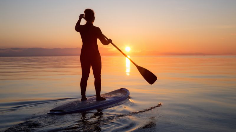 This image shows a person paddleboarding on a calm body of water during sunset. The person is silhouetted against the orange and pink hues of the sky, with the sun low on the horizon. The water is still, reflecting the colors of the sunset, and the person is standing on the paddleboard, holding a paddle as they glide across the water.