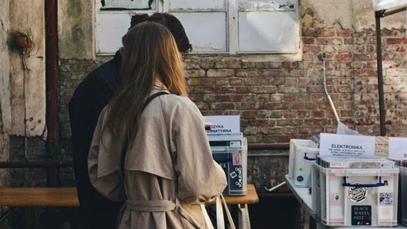 This image shows two people, one wearing a light-colored trench coat, browsing through a collection of vinyl records or CDs at an outdoor market. The individuals are facing away from the camera, and the setting features a rustic brick wall and an old window, giving the scene a vintage, urban atmosphere. 