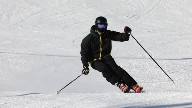 The image shows a skier dressed in a black jacket, helmet, and goggles carving down a snow-covered slope. 