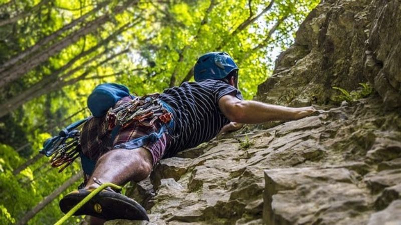 The image shows a rock climber scaling a vertical rock face in a lush forest setting. The climber, wearing a helmet, climbing gear, and harness, grips the rough surface of the rock while making their ascent.