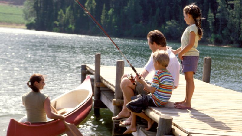 A family enjoying leisure time on a lakeside dock; children fishing, a woman sitting in a canoe, and lush green trees surrounding the calm lake.